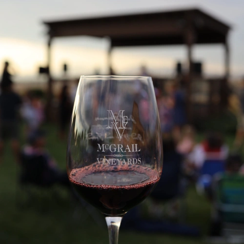 McGrail Vineyards wine glass filled with red wine at outdoor vineyard event with pergola and guests blurred in background.