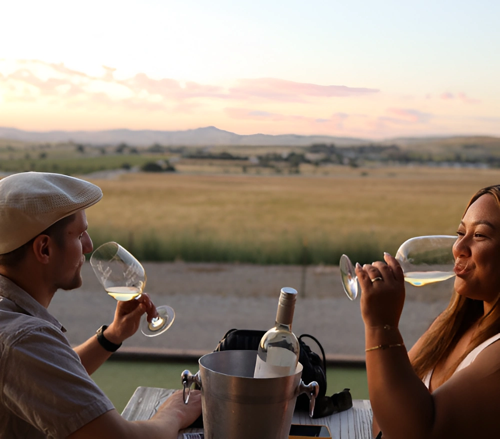 Two people holding wine glasses on a terrace with valley views and sunset sky, wine bucket on table. 