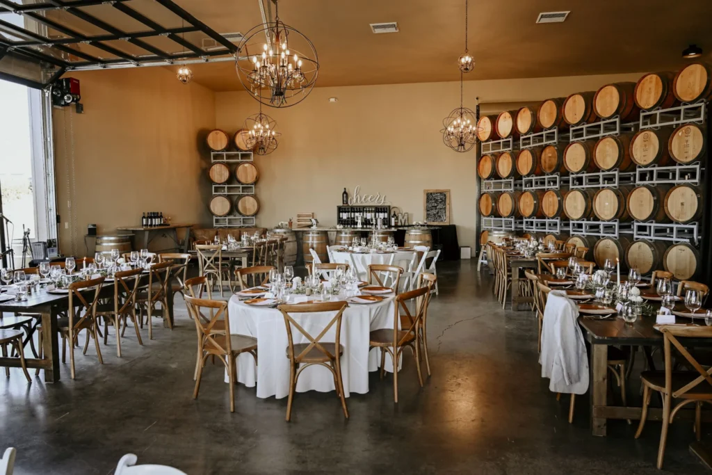 Wine barrel-lined event space with round dining tables, white linens, wooden chairs, and pendant lighting for private events.