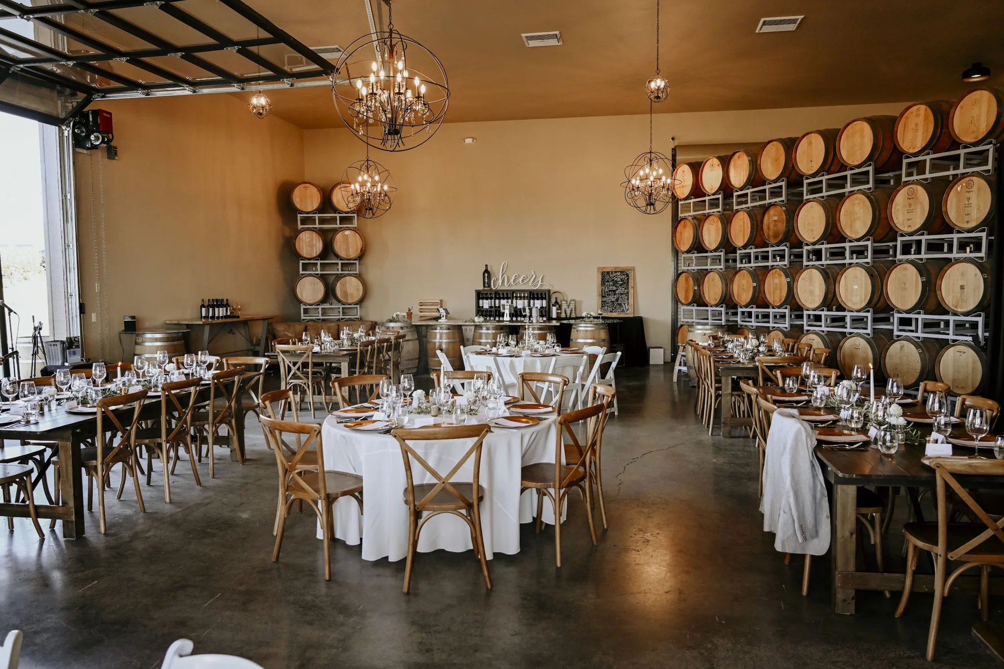 Wine barrel-lined event space with round dining tables, white linens, wooden chairs, and pendant lighting for private events.