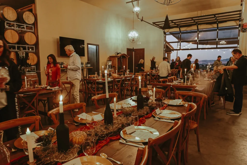 Winery dining event with set tables, wine bottles, and guests in a barrel-lined tasting room with vineyard views.
