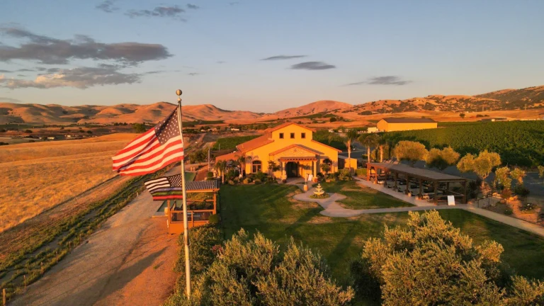 Yellow winery building with arches and American flag overlooking McGrail Vineyard's rolling terrain at sunset.