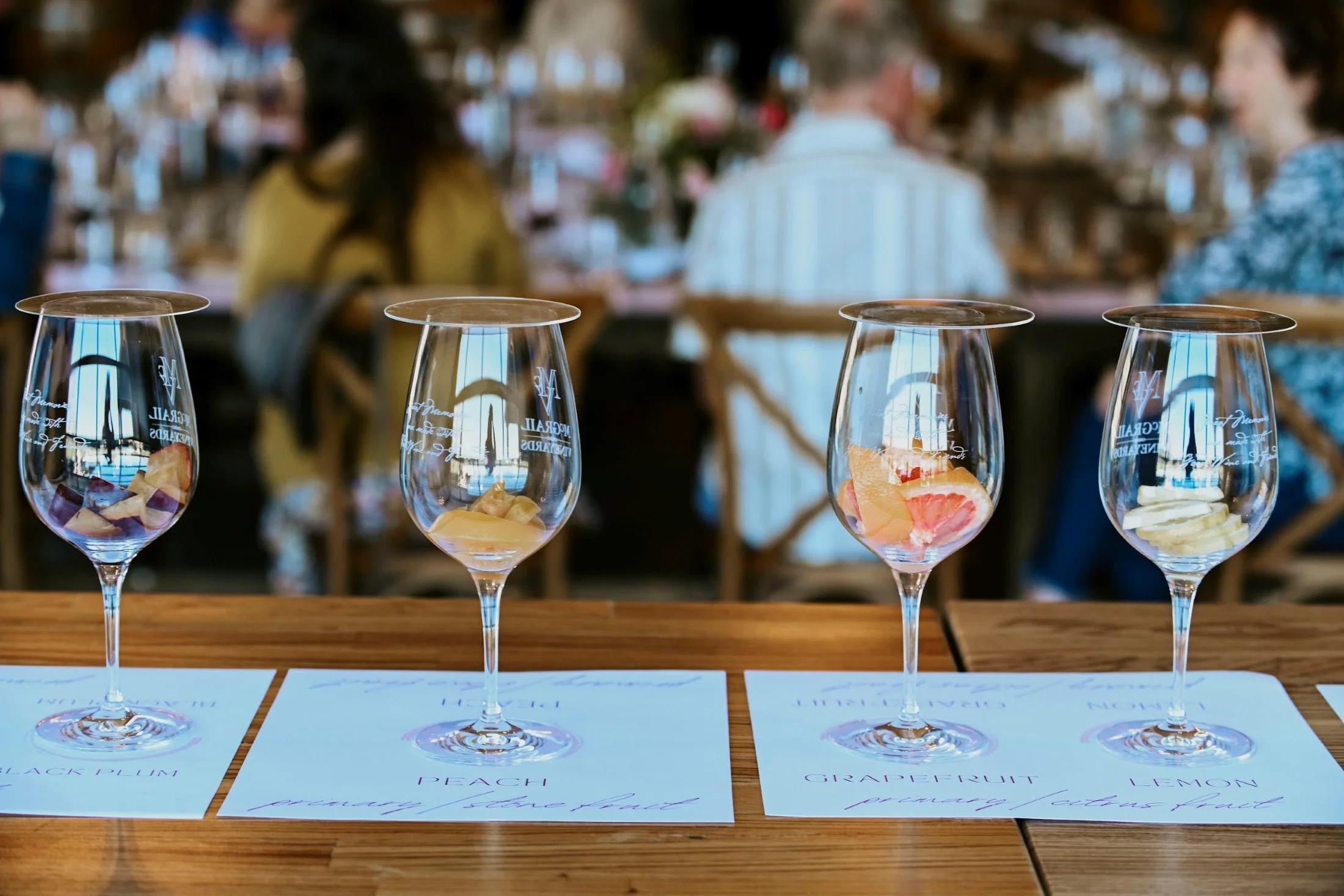 Four etched wine glasses with varying wine samples and ice arranged on a wooden bar with tasting cards.