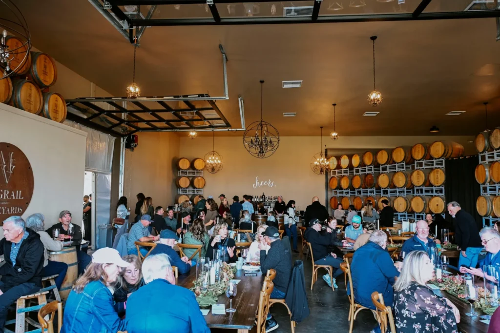 Guests at a wine release party in a vineyard barrel room with oak barrels lining the walls.