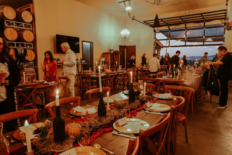 Winery dining event with set tables, wine bottles, and guests in a barrel-lined tasting room with vineyard views.