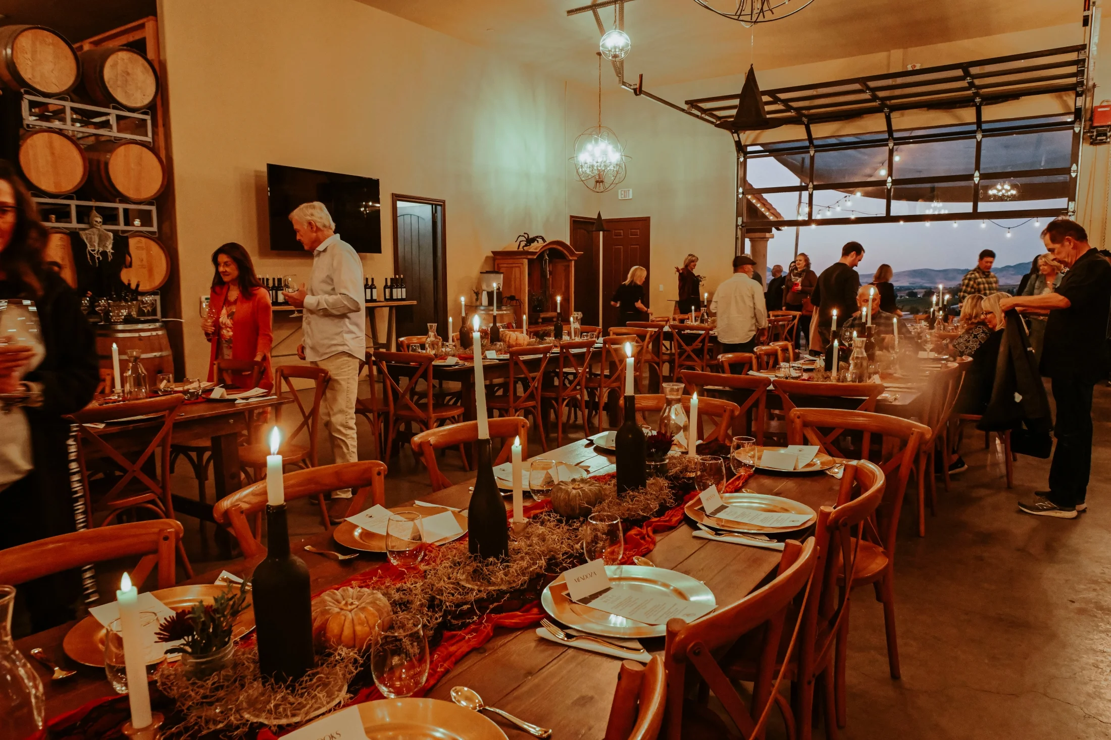 Winery dining event with set tables, wine bottles, and guests in a barrel-lined tasting room with vineyard views.