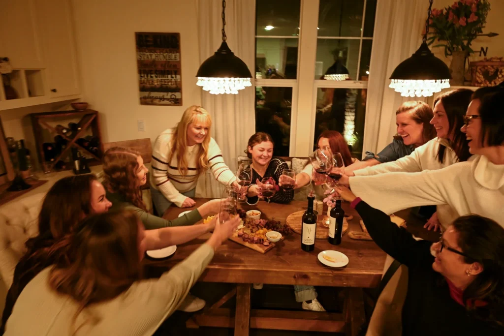 Women toasting with wine glasses around a wooden dining table during an indoor gathering.